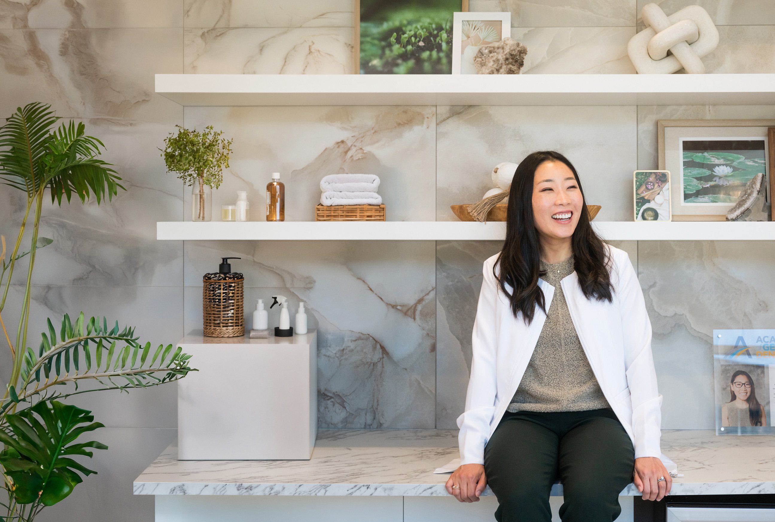 Dr. Erin Cho laughing, sitting on the counter under the shelf wall at Re·gen Smile Studio